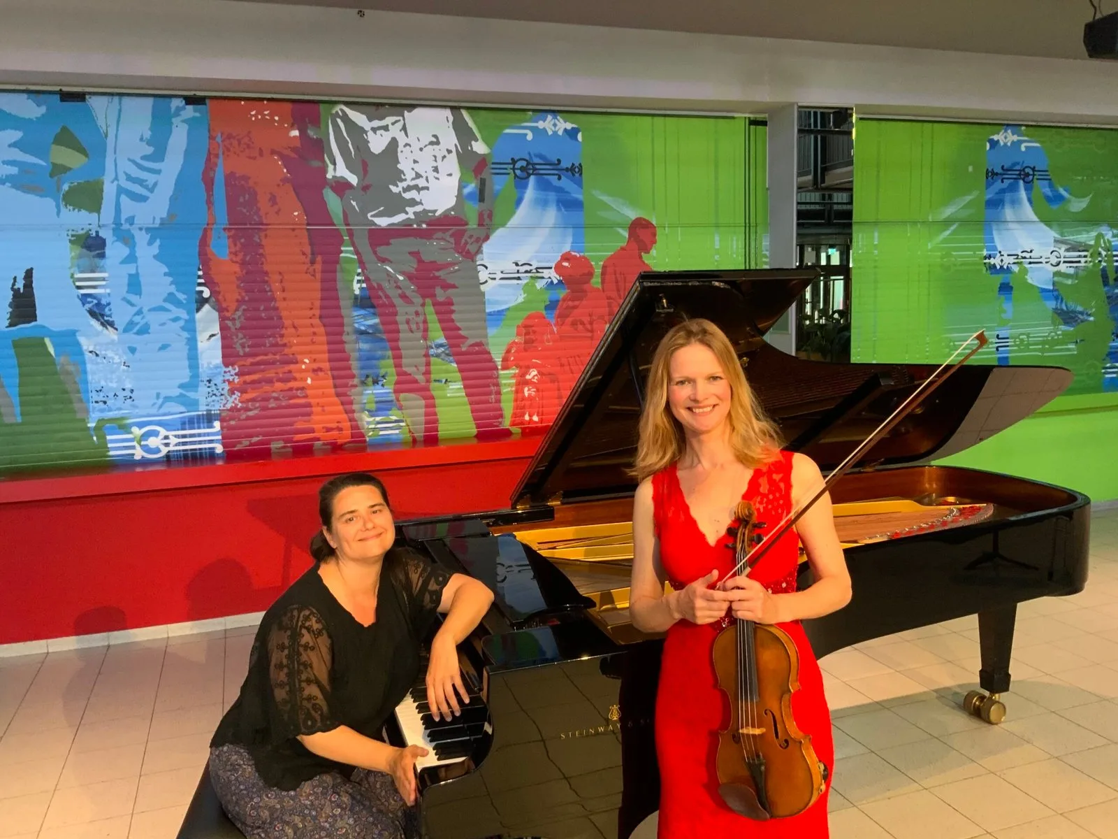 Evelina zit aan een vleugel naast een violiste die haar instrument vasthoudt